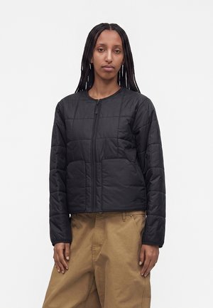 Persona con capelli lunghi e intrecciati che indossa una giacca nera trapuntata con cerniera e pantaloni larghi color sabbia, in piedi su uno sfondo bianco semplice.
