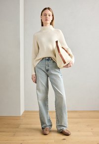 Beige ribbet genser, lyseblå høy midje jeans, brune slipsko, holder en lerretsveske med brun stropp. Bakgrunn av tregulv.