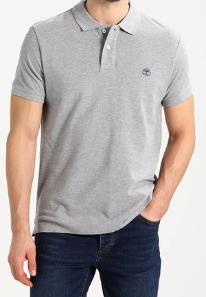 Homme portant un polo gris clair à manches courtes avec des boutons et un jean bleu foncé, main dans la poche, debout devant un fond blanc.