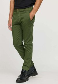 Chino olive avec une coupe slim, présentant une fermeture à bouton, des poches avant et des manchettes roulées, assortis à des bottes noires à lacets.