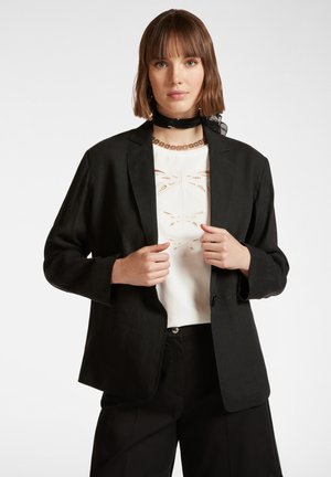 Schwarzer Blazer mit lockerer Passform, Ein-Knopf-Verschluss und leicht gerollten Ärmeln, getragen über einem weißen Top mit ausgeschnittenen Libellenmustern.