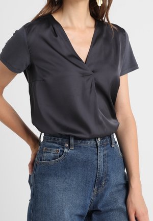 Woman wearing a short-sleeve black satin V-neck blouse tucked into high-waisted blue jeans with a hand resting on her hip.