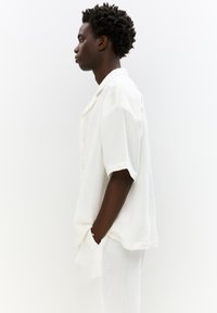 Camicia bianca a maniche corte con colletto, leggere pieghe e vestibilità comoda, abbinata a pantaloni coordinati, mostrata di lato.