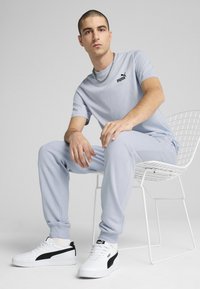 Un tricou bleu deschis și pantaloni joggeri asortați dintr-un material moale. Logo-ul negru Puma pe tricou. Adidași albi cu accente negre. Colier din lanț metalic.