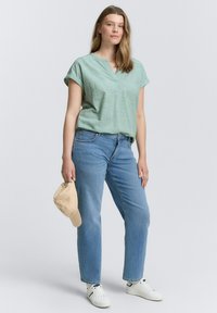 Light green short-sleeve blouse with a V-neck, paired with medium blue straight-leg jeans and white sneakers. Holding a beige cap.