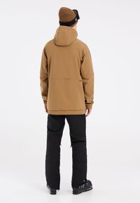 Giacca a cappuccio marrone con una superficie liscia, abbinata a pantaloni neri e scarponi da sci con fibbie, mostrata dal retro su uno sfondo chiaro.