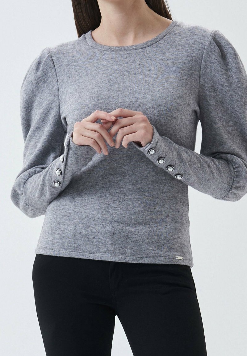 Maglione grigio con maniche a sbuffo, caratterizzato da bottoni decorativi ai polsini e una texture morbida. Indossato con pantaloni neri aderenti.