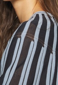 Bluzka w paski z pionowymi liniami w kolorze czarnym i jasnoniebieskim. Teksturowana tkanina o luźnym kroju i marszczeniu przy dekolcie.