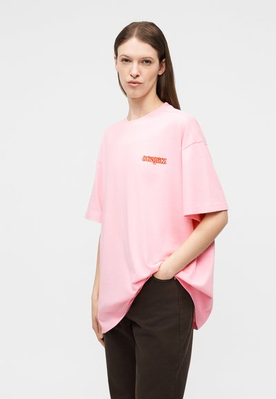 Γυναίκα που φοράει ένα άνετο ροζ T-shirt με κόκκινα γράμματα στο στήθος και σκούρο παντελόνι, στέκεται με το ένα χέρι στην τσέπη μπροστά σε απλό φόντο.