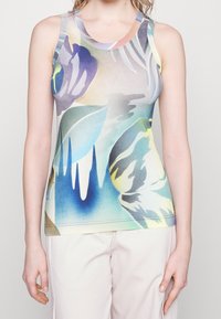 Kobieta w bezrękawniku z abstrakcyjnym nadrukiem w odcieniach niebieskiego, zielonego, żółtego i fioletowego, w zestawieniu z jasnymi spodniami na jednolitym tle.