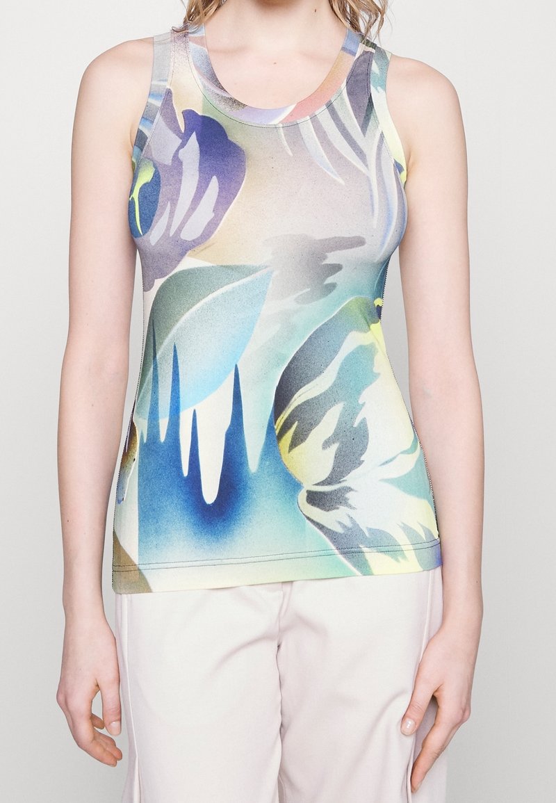 Kobieta w bezrękawniku z abstrakcyjnym nadrukiem w odcieniach niebieskiego, zielonego, żółtego i fioletowego, w zestawieniu z jasnymi spodniami na jednolitym tle.