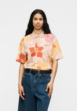 Žena s dugom tamnom kosom u višeobojnoj polo majici s cvjetnim uzorkom i plavim visokim trapericama s crnim remenom, stoji na bijeloj pozadini.