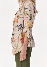 Kvinde iført en beige bluse med blomsterprint og oprullede ærmer, samt grønne bukser, vist fra siden mod en ensfarvet baggrund.
