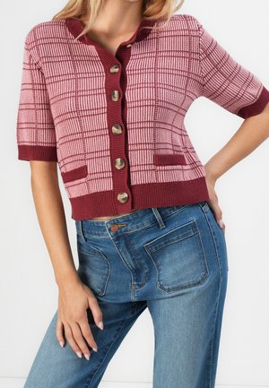 Femme portant un cardigan à manches courtes à carreaux rose et bordeaux, associé à un jean bleu taille haute.