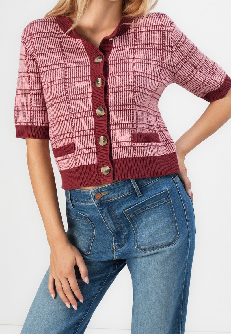 Femme portant un cardigan à manches courtes à carreaux rose et bordeaux, associé à un jean bleu taille haute.