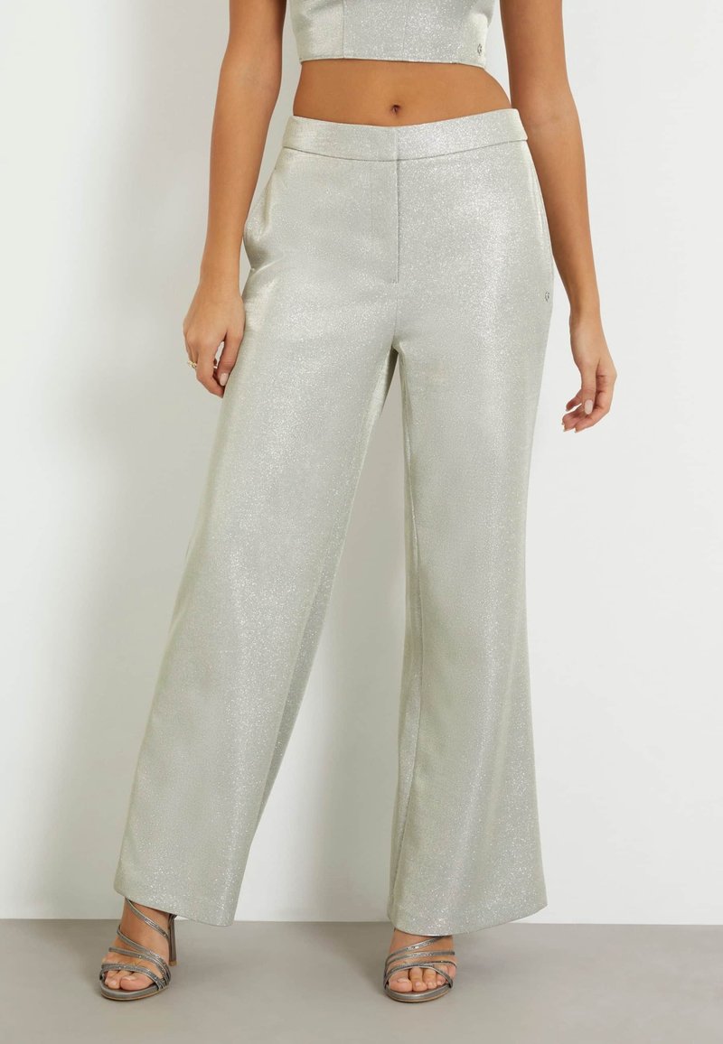Pantalons larges argentés en tissu brillant et texturé. Dotés d'une taille lisse et de poches sur les côtés. Associés à des sandales à talons métalliques.