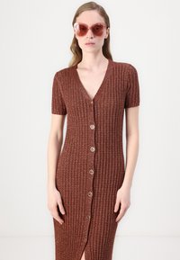 Knieumspielendes, figurbetontes, kurzärmliges Kleid aus strukturiertem braunem Strick mit V-Ausschnitt und goldenen Knöpfen als Frontverschluss. Große transparente Sonnenbrille.