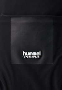 Juodas sportinis audinys su tekstūru ir permatomu lopu. Iškilios baltos spalvos logotipas skelbia "hummel SPORTSWEAR".