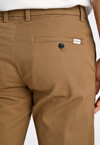 Kaki byxor gjorda av en bomullsmix, med en bakficka som har en svart knapp och en liten varumärkesetikett sydd ovanför.