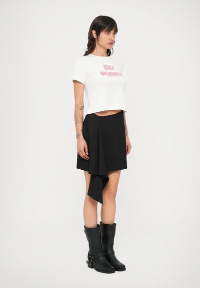 Valge lühem T-särk, millel on roosades karvastes tähtedes kiri "SKI BUNNY", kombineeritud musta asümmeetrilise seeliku ja põlvekõrguste mustade nahk saabastega.