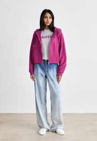 Violet zip-up džemperis virš pilkos marškinėlių su "BOSS" logotipu, derinamas su plačiomis šviesiai mėlynomis džinsų kelnėmis ir baltais sportbačiais. Vienoda fonas.