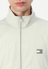 Lys grønn zip-up jakke med høy krage, laget av et glatt stoff. Har et logo-patch på brystet og sølv glidelåsdetaljer.