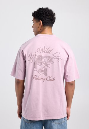 Man draagt lichtroze T-shirt met tekst "The Wild Life Fishing Club" en een visafbeelding op de achterkant, gecombineerd met een blauwe spijkerbroek.