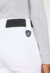 Fehér nadrág fekete elasztikus derékpánttal, amelyen az "ARMANI" felirat és egy EA7 logós címke található a hátsó zsebén. Szürke hosszú ujjú felső látható.