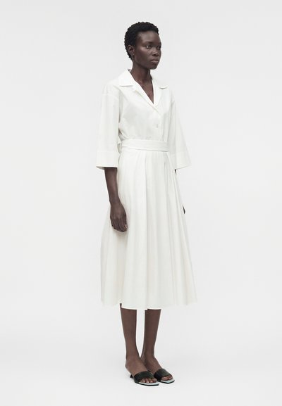 Γυναίκα με λευκό, κουμπωτό φόρεμα μεσαίου μήκους με γιακά και φαρδιά μανίκια, φοράει μαύρα ανοιχτά πέδιλα με χαμηλό τακούνι, στέκεται σε λευκό φόντο.