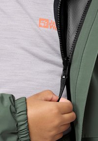 Grön jacka med elastiska ärmslut, blixtlåsförsedd front och grå skjorta under med en orange logotyp. Närbild av hand som håller i blixtlåsdragningen.