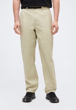 Άνδρας που φοράει μπεζ παντελόνι με ελαστική γραμμή στη μέση και μαύρη ζώνη, σε συνδυασμό με μαύρα αθλητικά παπούτσια, στέκεται μπροστά σε απλό φόντο.