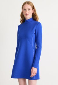 Modra dolga obleka z dolgimi rokavi iz gladkega blaga, ki ima visok ovratnik, ukrivljene šive in majhen logotip na robu.