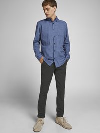 Plava dugosleeve košulja s gumbima i džepom na prsima, u kombinaciji s tamnim slim-fit hlačama i bež cipelama s vezicama, stoji protiv svijetle pozadine.