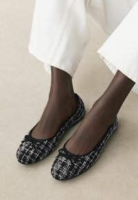 Schwarz-weiße Tweed-Ballerinas mit einer abgerundeten Spitze, verziert mit einer schwarzen Schleife, die ein strukturiertes Muster auf dem Obermaterial und eine flache Sohle aufweisen.
