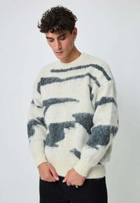 Pull en laine blanche avec motifs abstraits noirs et gris, manches longues, poignets et ourlet côtelés, texture douce, coupe légèrement oversize.