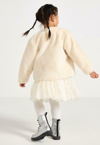Fleece-Creme-Jacke, schichtiges weißes Kleid mit Rüschen, strukturierte weiße Strumpfhosen und silberne Schnürstiefel mit schwarzen Akzenten.