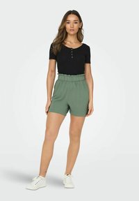 Top preto canelado de manga curta com detalhes em botão combinado com calções verdes de cintura elástica. Modelo usa ténis brancos. Fundo liso.