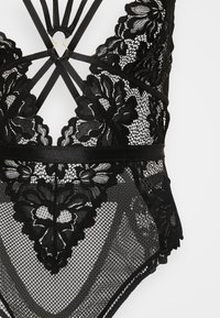 Μαύρο κορμάκι από δαντέλα με ανθικά σχέδια, ρυθμιζόμενους ιμάντες, διαφάνειες από δίχτυ και μεταλλικό κρίκο στο κέντρο.