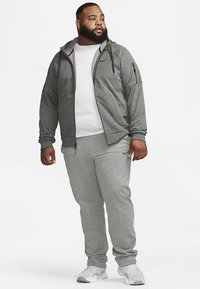 Casaco desportivo cinzento com capuz e bolsos com fecho, combinado com calças de treino cinzentas a condizer. O material parece macio com acabamento texturizado.