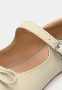 Δερμάτινο παπούτσι σε κρεμ απόχρωση με στρογγυλεμένη μύτη, αποσπώμενο λουράκι και μικρό ασημένιο πόρπη. Το εσωτερικό διαθέτει απαλό μπεζ φόδρα.