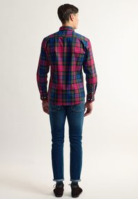 Camisa de cuadros de manga larga en rojo, azul y verde, con cuello puntiagudo y puños abotonados, combinada con vaqueros ajustados azules y zapatos marrones.