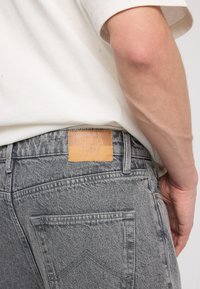 Pantalones vaqueros de mezclilla gris con un acabado texturizado, que presentan un parche de cuero marrón claro etiquetado como "AMERICA TODAY" en la cintura y en los bolsillos traseros.