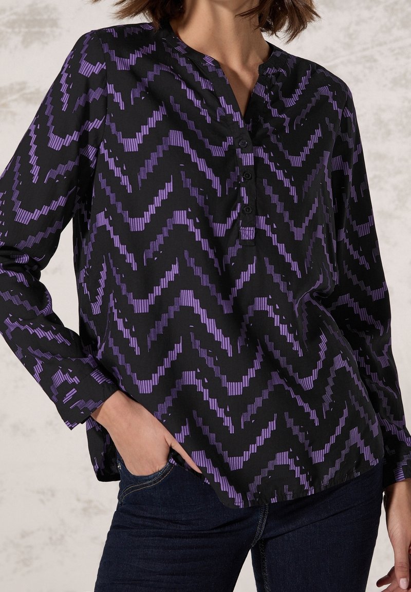 Blouse noire avec un motif en zigzag violet. Dotée d'un col en V, d'une fermeture à boutons et de manches longues. Confectionnée en tissu lisse.
