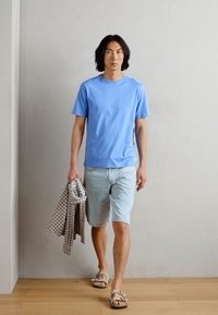Lyseblå t-skjorte med korte ermer, lyse denimshorts og sandaler. Holder en rutete skjorte. Enkelt design på en menneskelig figur mot en nøytral bakgrunn.