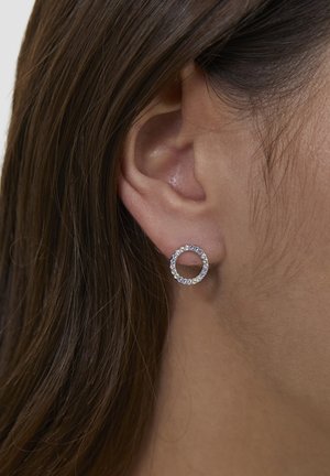 Runde silberne Ohrringe mit einem kreisförmigen Design, verziert mit kleinen klaren Kristallen am Rand, vor einem Haarhintergrund.