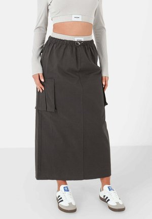 Langer, dunkelgrauer Cargo-Rock mit elastischem Bund, seitlichen Taschen und gerader Silhouette. Kombiniert mit weißen Sneakers mit schwarzen Streifen.