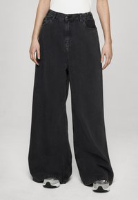 Černé široké džíny z denimu s vysokým pasem, vybavené poutky na pásek, předními kapsami a volným střihem. Nositelná se šedými teniskami.