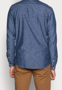 Mężczyzna ubrany w niebieską koszulę na guziki z długim rękawem i brązowe spodnie, widziany od tyłu na jednolitym tle.