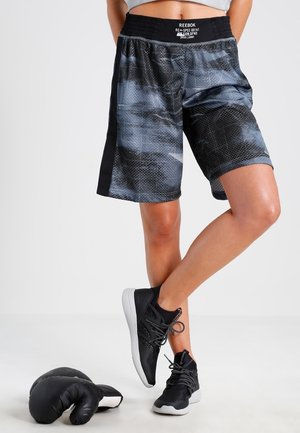 Persona che indossa pantaloncini sportivi Reebok mimetici neri e grigi e scarpe da ginnastica nere, in piedi con una gamba incrociata sull'altra accanto a guantoni da boxe neri.