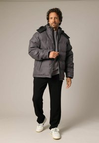 Grijze gewatteerde jas met hoge kraag en afneembare capuchon, gecombineerd met een zwarte broek en witte sneakers, tegen een neutrale achtergrond.